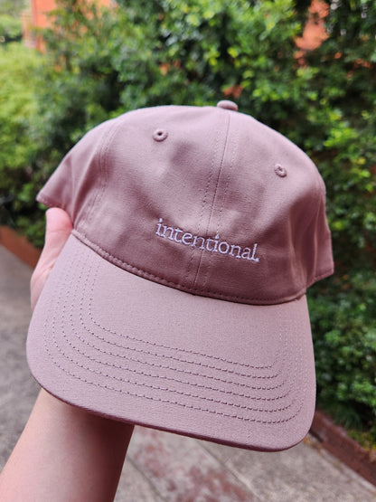Pink cap held by a hand with 'Intentional' embroidery, outdoor background