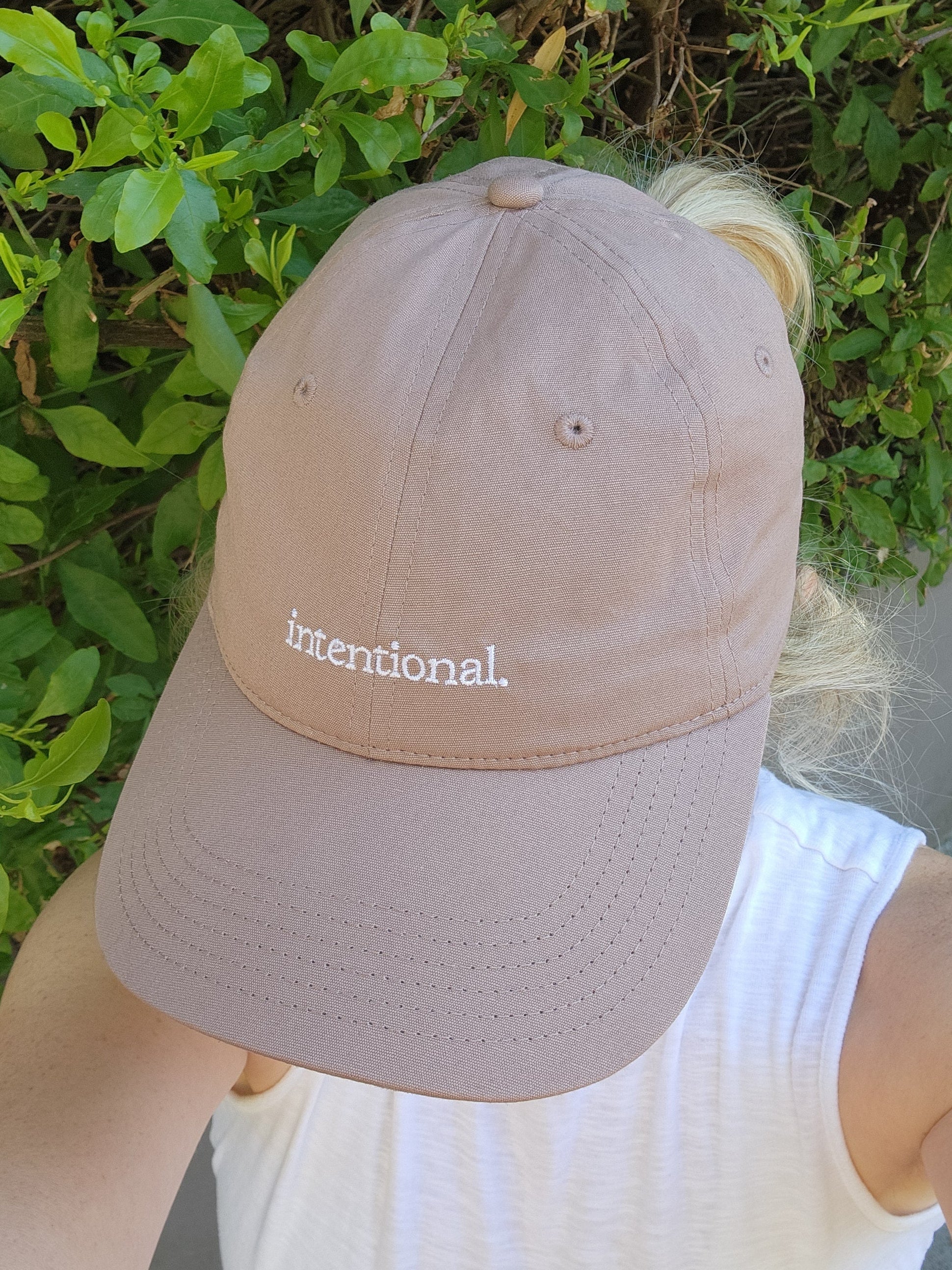 Dusty Pink cap with 'intentional' text embroidered on it. Worn by a person against a green leafy background.