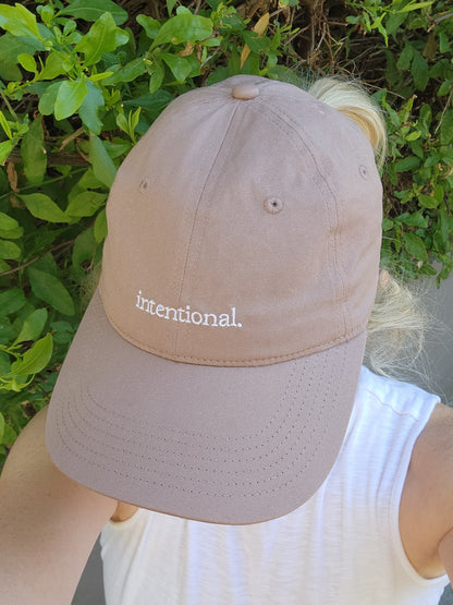 Dusty Pink cap with 'intentional' text embroidered on it. Worn by a person against a green leafy background.