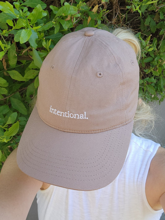 Dusty Pink cap with 'intentional' text embroidered on it. Worn by a person against a green leafy background.